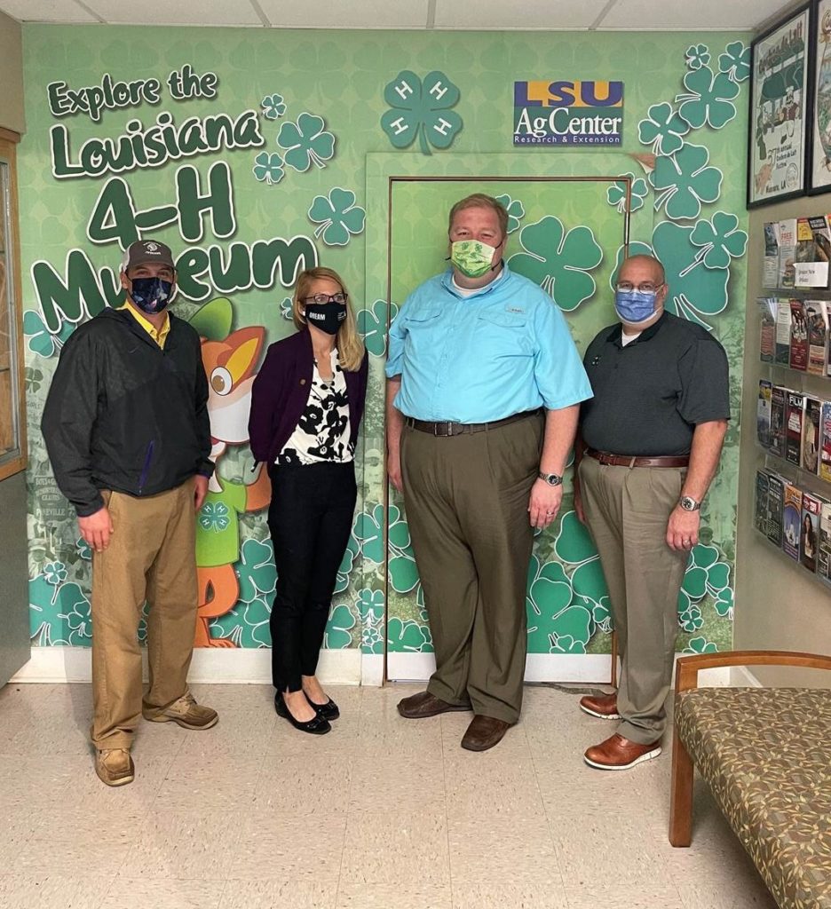 Mr. Justin Crowe standing with 3 members of the Louisiana 4-H staff against a clover mural that says "Explore the Louisiana 4-H Museum."