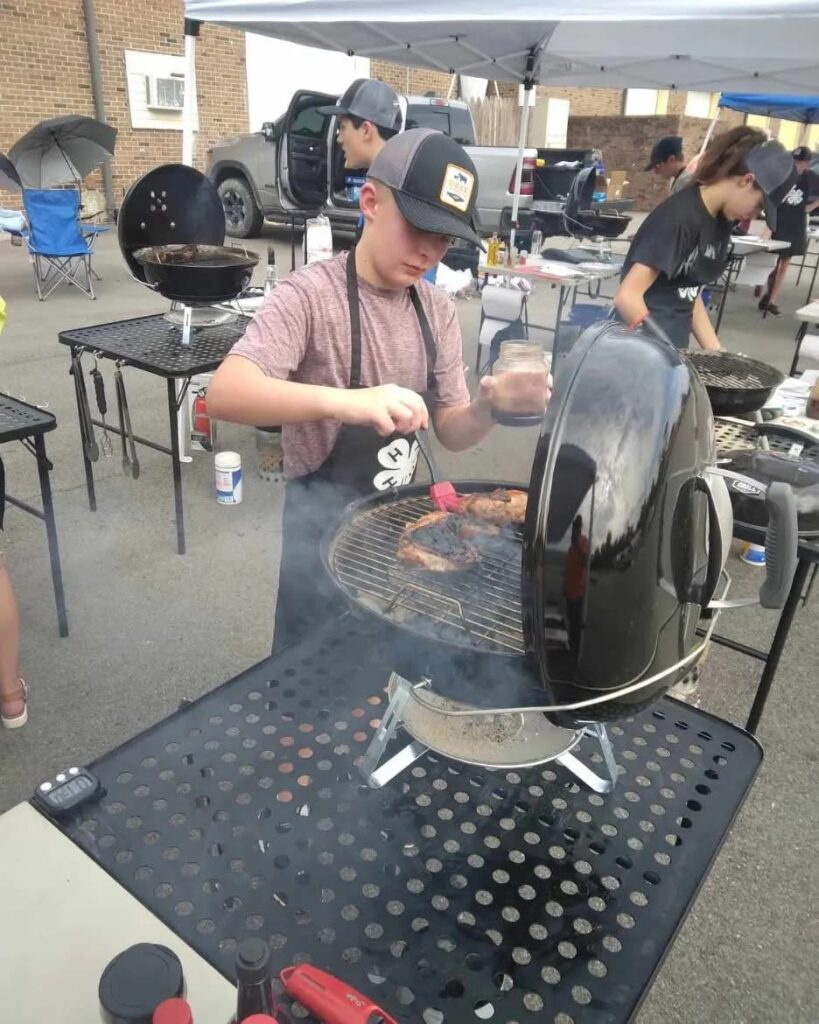 youth basting meat on a table top grill with smoke coming from the grill.